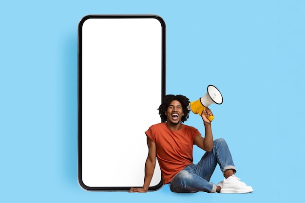 Happy black guy with loudspeaker sitting by cell phone mockup