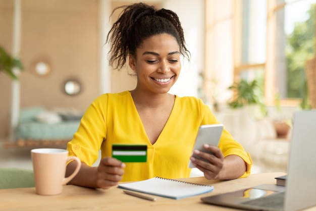 Happy black lady buying things online using smartphone and credit card shopping in internet while sitting at home