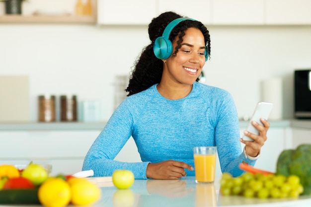 Happy black lady in sportswear using smartphone and wireless headphones listening music in kitchen