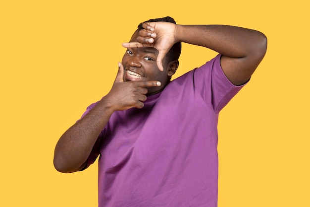 Happy black man making picture frame with fingers yellow background