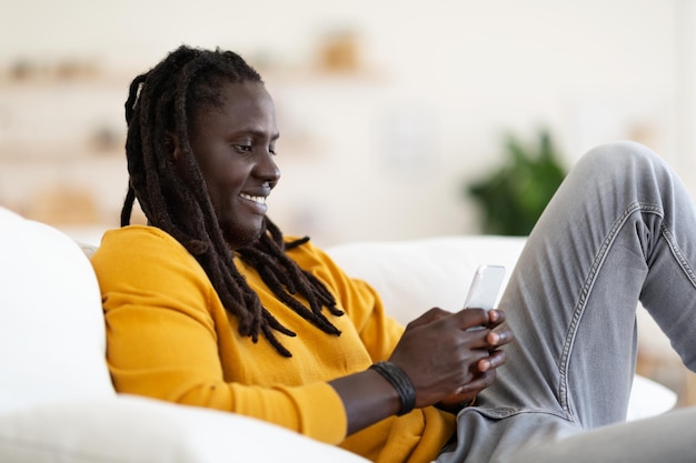 Happy black man messaging on smartphone while relaxing on couch at home