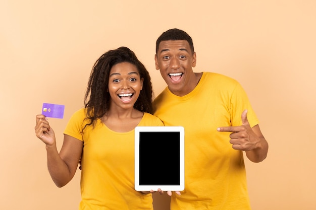 Happy black man pointing at empty tablet screen his girlfriend holding credit card on peach studio background mockup