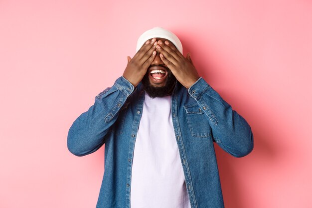 Happy Black man waiting for surprise, cover eyes and smiling, playing peekaboo, standing over pink background.