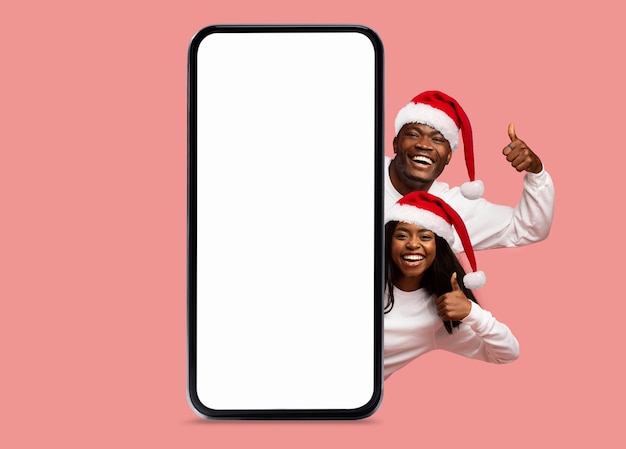 Happy black millennial woman and man in Santa Claus hats show thumbs up with big smartphone