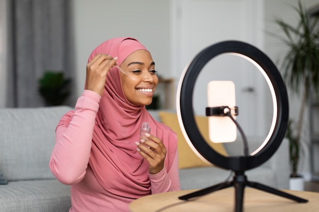 Happy black muslim female beauty blogger making video on smartphone set on ring lamp showing new facial serum