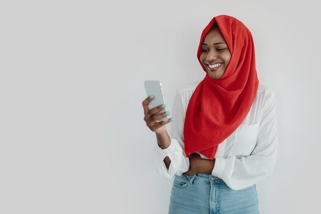 Happy black muslim woman in headscarf using smartphone texting or browsing internet light wall free space