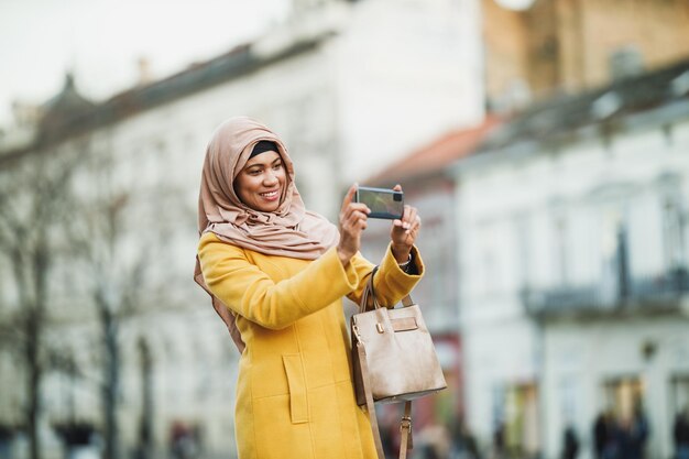 Happy black Muslim woman wearing a hijab and taking photos by her smartphone while standing in urban environment.