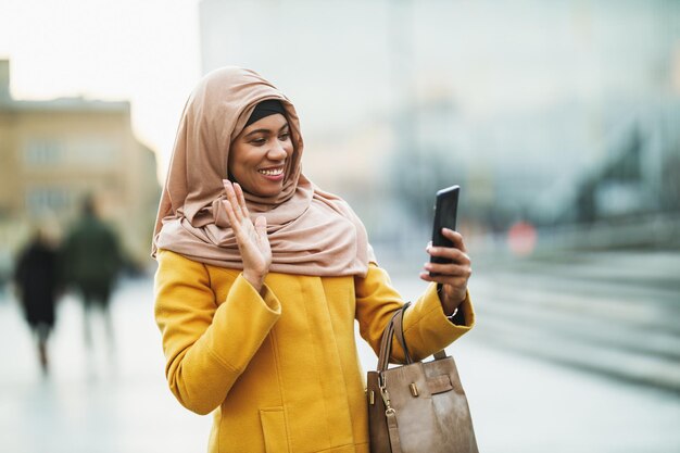 Happy black Muslim woman wearing a hijab and using her smartphone while walking downtown.