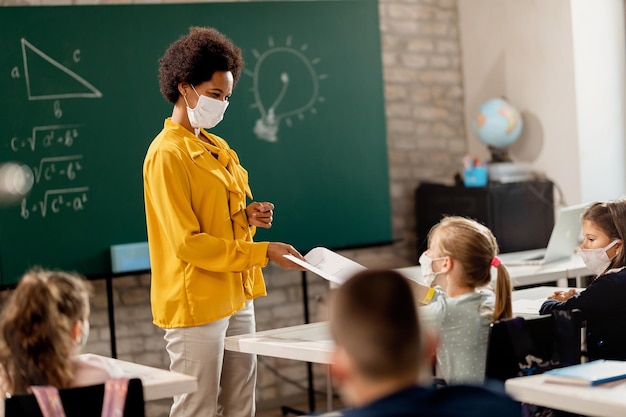 Happy black teacher and her students wearing protective face mask in the classroom. Teacher is giving them their test results.