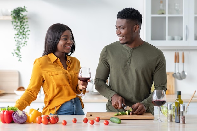 Happy black woman drinking wine while her husband cooking salad in kitchen