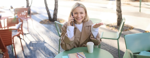 Happy blond female model sitting outdoors in cafe answer phone call having conversation on