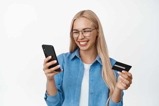 Happy blond woman smiling wearing glasses looking at mobile phone screen while paying for online order holding credit card standing over white background