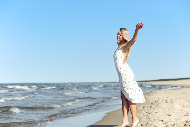 Happy blonde beautiful woman on the ocean beach standing in a white summer dress, open arms.