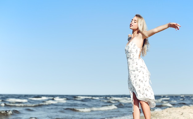 Happy blonde beautiful woman on the ocean beach standing in a white summer dress, open arms