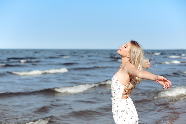 Happy blonde beautiful woman on the ocean beach standing in a white summer dress, open arms.