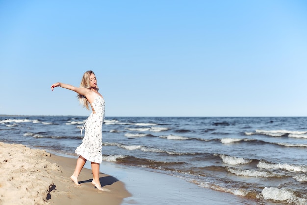 Happy blonde beautiful woman on the ocean beach standing in a white summer dress, open arms