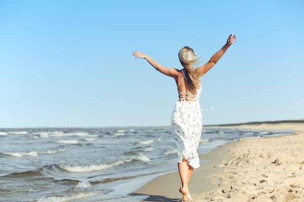 Happy blonde beautiful woman on the ocean beach standing in a white summer dress, open arms.