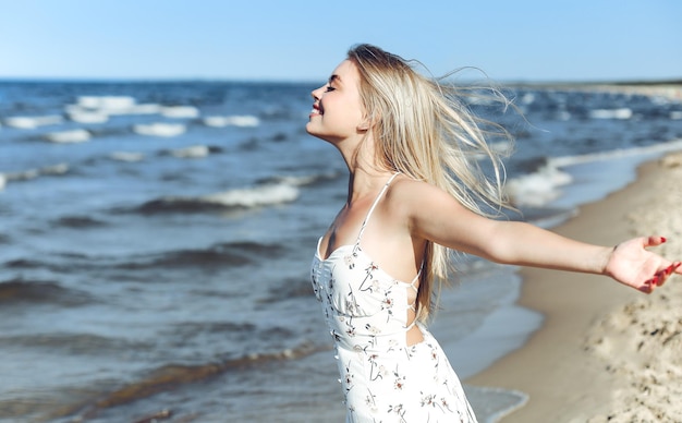 Happy blonde beautiful woman on the ocean beach standing in a white summer dress, raising hands.