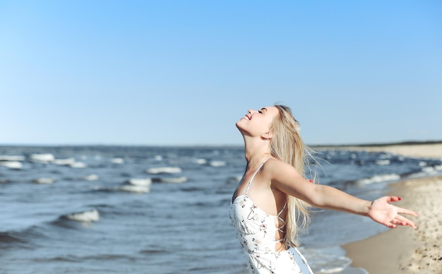 Happy blonde beautiful woman on the ocean beach standing in a white summer dress, raising hands.