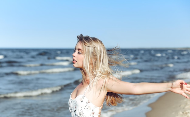 Happy blonde beautiful woman on the ocean beach standing in a white summer dress, raising hands