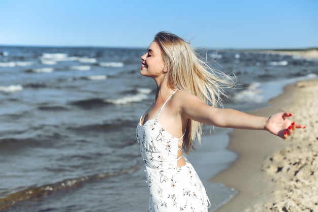 Happy blonde beautiful woman on the ocean beach standing in a white summer dress, raising hands.