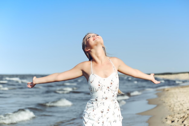 Happy blonde beautiful woman on the ocean beach standing in a white summer dress, raising hands.