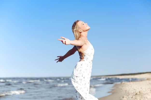 Happy blonde beautiful woman on the ocean beach standing in a white summer dress, raising hands.