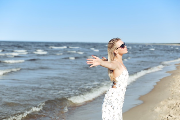 Happy blonde beautiful woman on the ocean beach standing in a white summer dress and sun glasses, open arms.