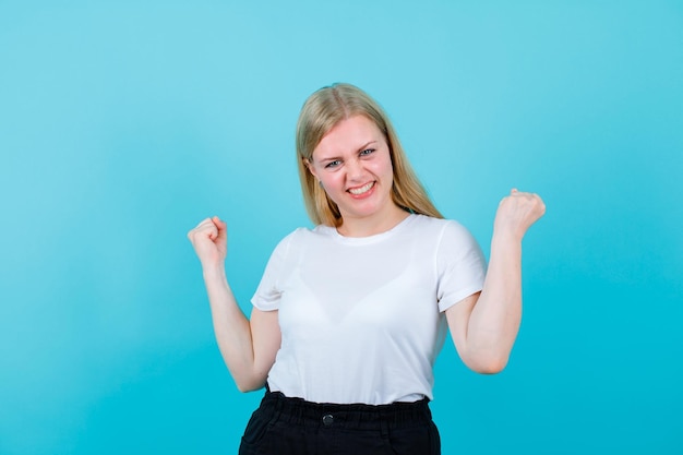 Happy blonde girl is raising up her fists on blue background