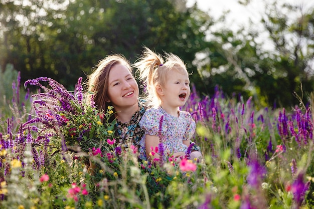 Happy blonde mom and daughter in a summer blooming field with a bouquet of wild flowers in their hands