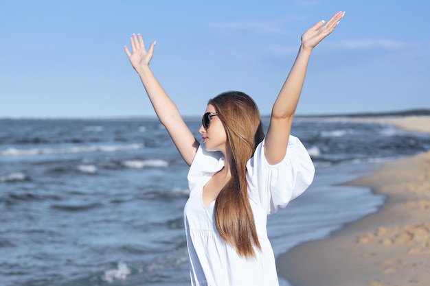 Photo happy blonde woman is on the ocean beach in a white dress and sunglasses raising hands