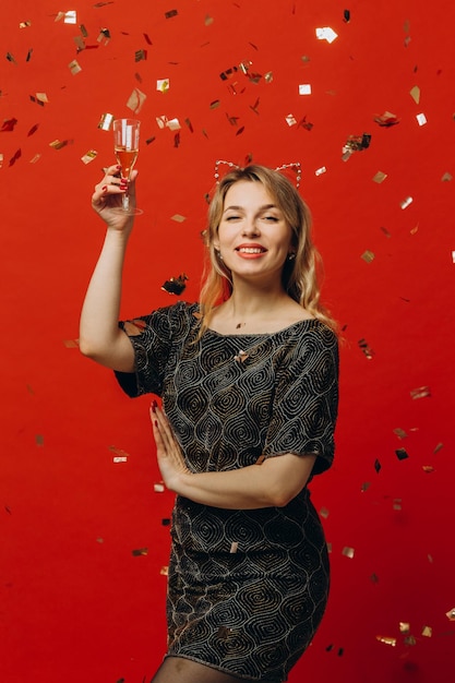 Happy blonde woman in shiny fashion clothes is happy holding glass of champagne and posing with confetti on red background