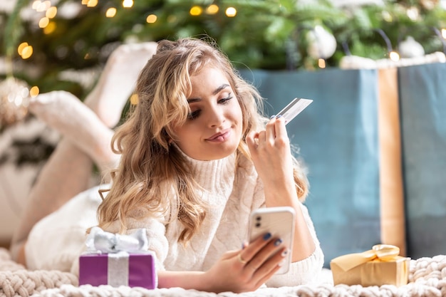 Happy blonde woman using smartphone and credit card shopping online lying on a blanket near gifts and Christmas tree at home