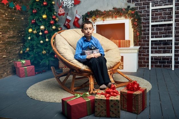 Happy boy in a comfortable chair near the Christmas tree by the fireplace with got a lot of gifts.