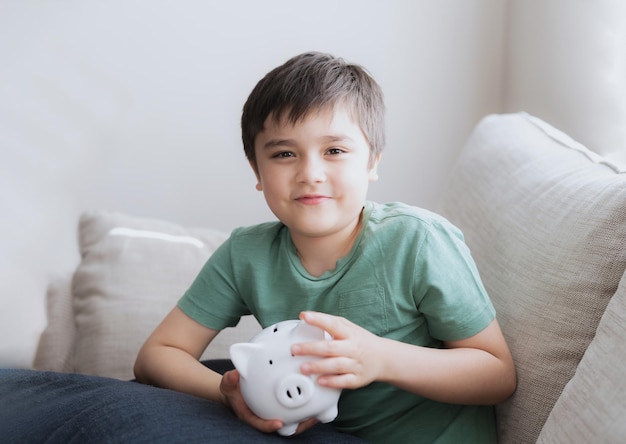 Happy boy holding piggy bank with smiling face Indoor portrait of a cheerful child showing money saving boxSchool kid Learning financial responsibility and planning about saving for future concept