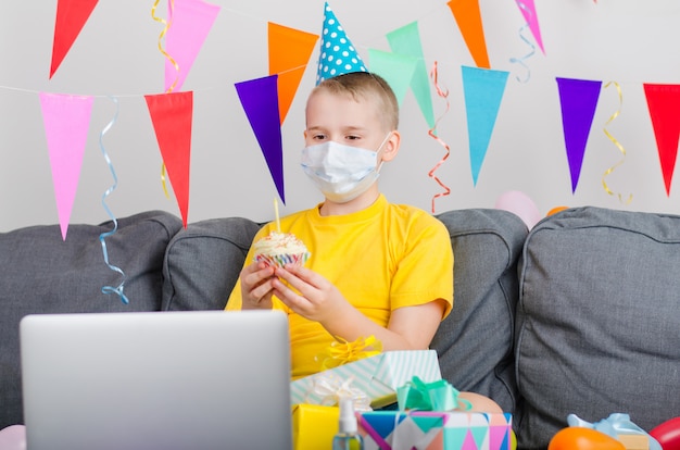 Happy boy in medicine face mask celebrates birthday by video call to laptop