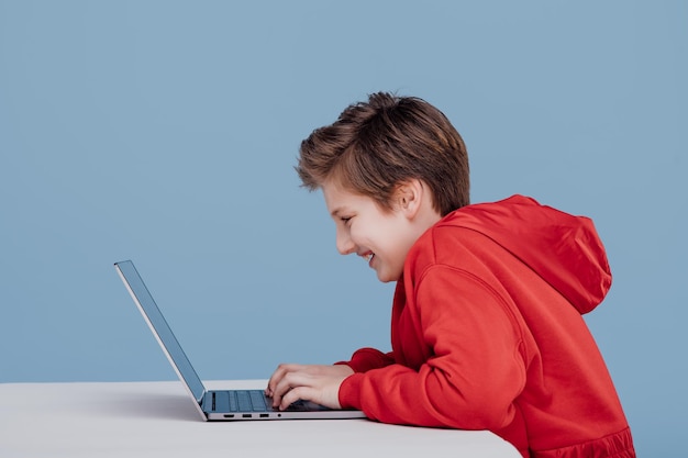 Happy boy n red sweatshirt playing on laptop iisolated blue background profile view