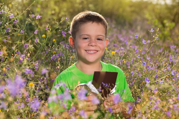 Happy boy in nature, with a bar of chocolate in his hands, surrounded by wildflowers