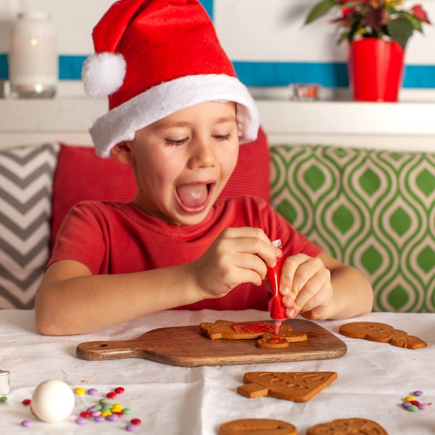 A happy boy in a santa claus hat decorates ginger cookies in the kitchen christmas lights