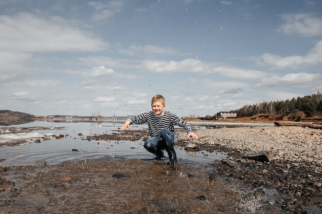 A happy boy in a striped vest and rubber boots on the bank of the river splashes water merrily. Outdoor recreation.