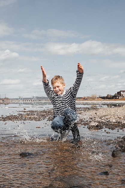 A happy boy in a striped vest and rubber boots on the bank of the river splashes water merrily. Outdoor recreation.