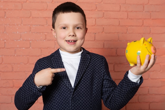 happy boy in suit holding a yellow piggy bank on brick background