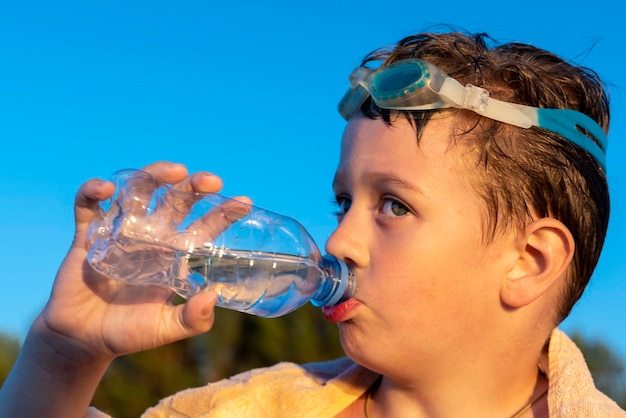 Happy boy in swimming glasses drinks water from a bottle on the beach