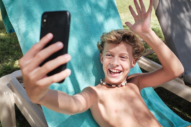 Happy boy with blond hair sitting on blue blanket and looking at screen of mobile phone while laughing with opened mouth and saying hello with raised hand fingers