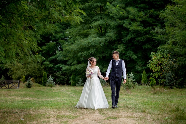 Happy bride and groom holding hands and walking in garden wedding day. Back view of charming stylish newlyweds holding hands while walking in park forest, happy marriage moments