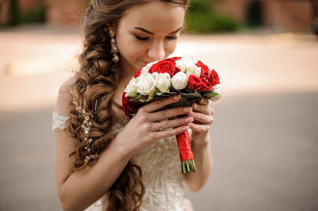 Happy bride in a wedding dress with a braid hairstyle sniffing a bouquet of red and white roses