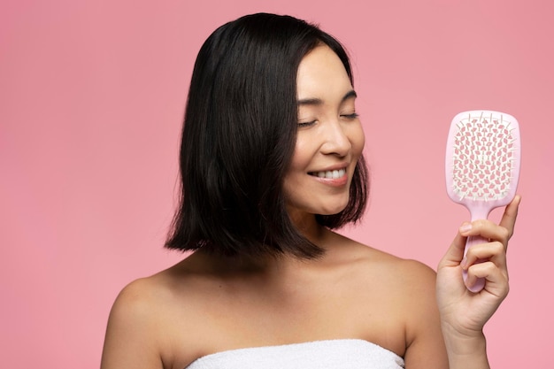 Happy brunette asian woman with silky hair holding modern hair brush isolated