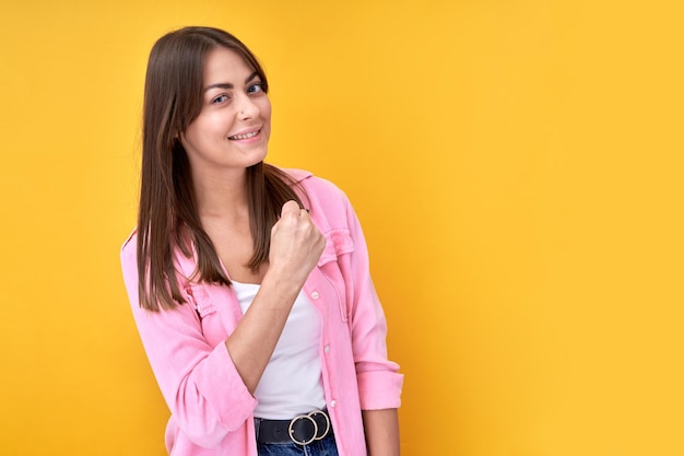 Happy brunette girl celebrating victory rejoices clenching fists isolated on yellow background