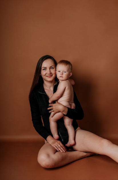 A happy brunette mom in a black bodysuit sits with her baby daughter on a brown background with a place for text