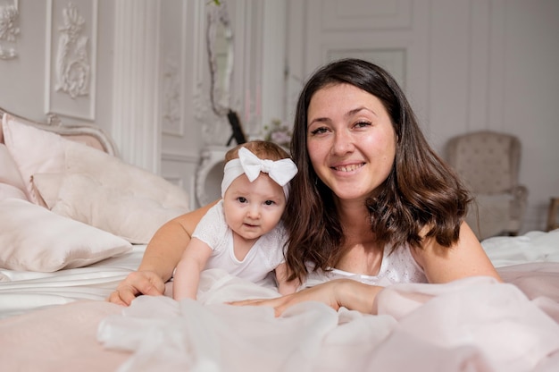 Happy brunette mom lies on the bed with her baby daughter and looks at the camera Single mother